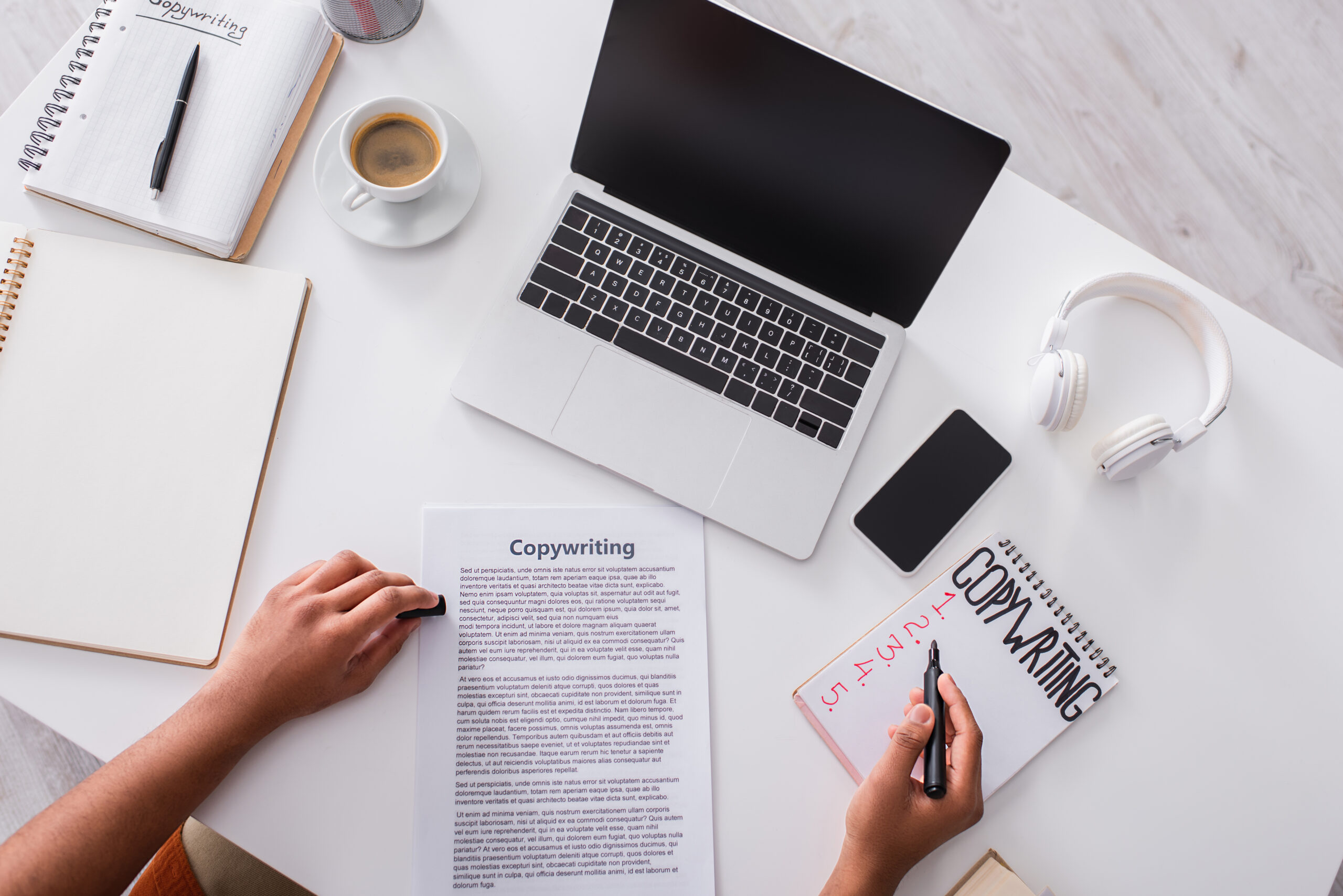 A person working on a copywriting project at a desk with a laptop, notebook, coffee cup, smartphone, and headphones.