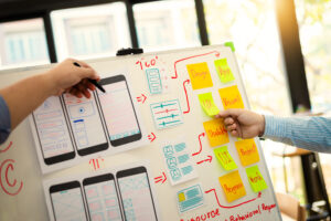 Hands pointing at a whiteboard with sketches of mobile app designs and sticky notes labeled with UI/UX, Visual, Usability, and Design.