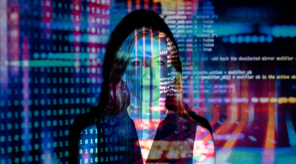 A woman standing in a dark room is illuminated by colorful, projected digital code and data patterns, her face partially obscured by the overlay of the code.