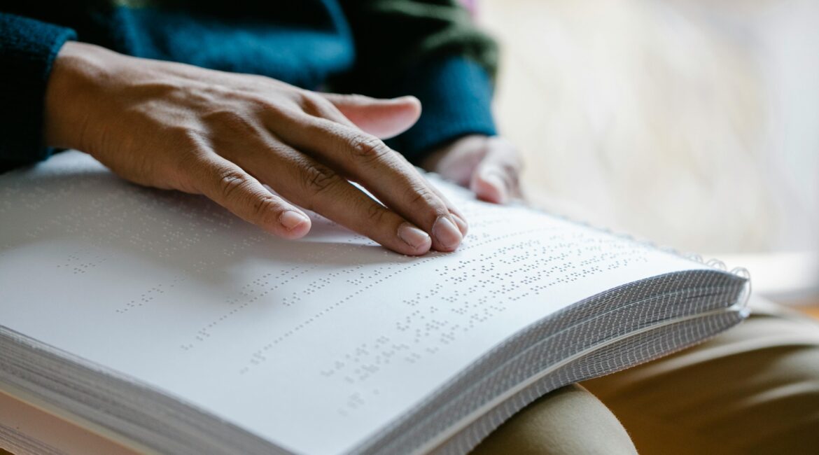 A close-up of a person's hand reading braille text on a large, spiral-bound book, highlighting their fingers as they move across the raised dots.