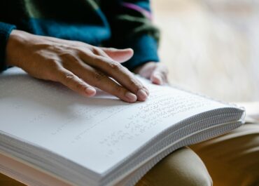 A close-up of a person's hand reading braille text on a large, spiral-bound book, highlighting their fingers as they move across the raised dots.