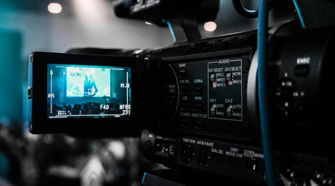 A close-up view of a professional video camera recording a live event, with the display screen showing the audience and stage in the background.