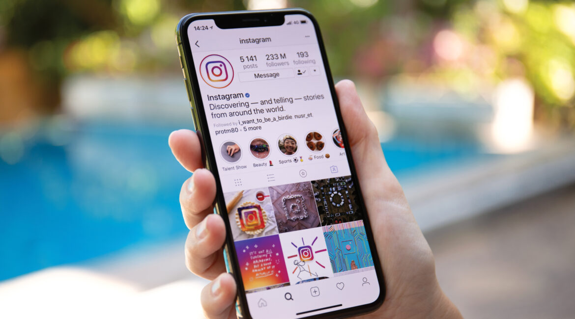 A person holding a smartphone displaying the Instagram profile page, with a background of a blurred swimming pool and greenery.