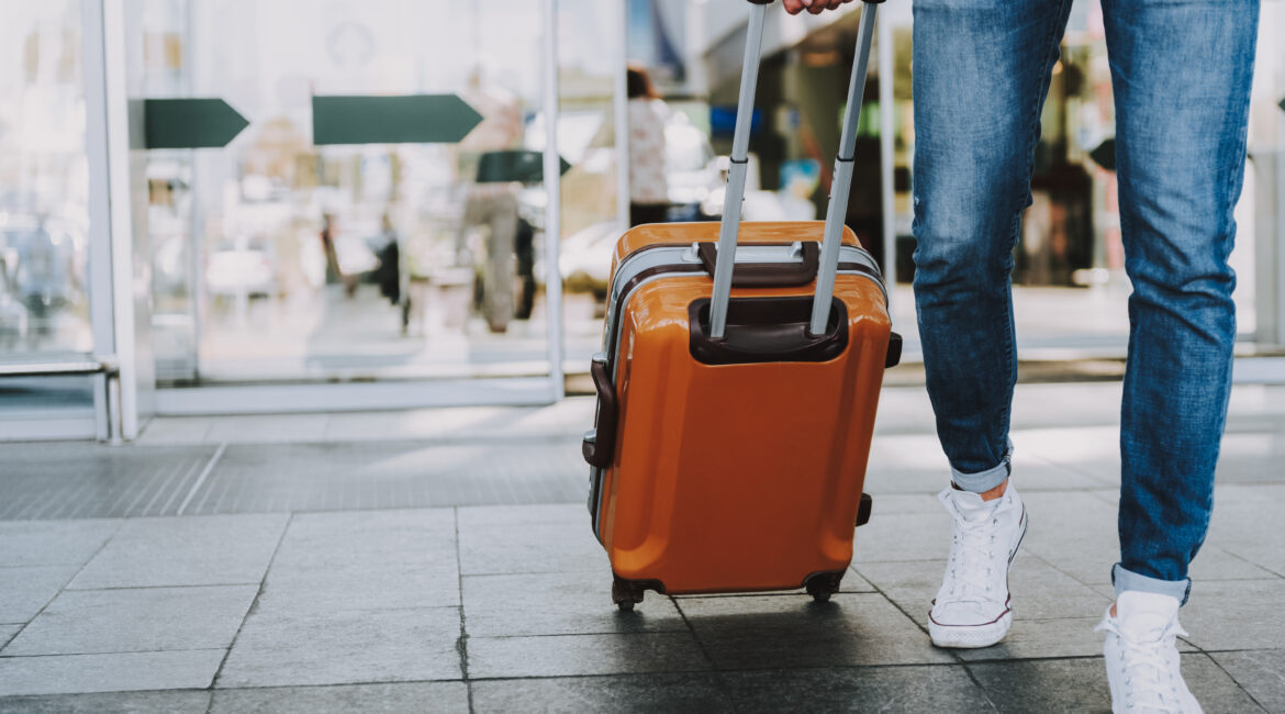 A traveler pulling an orange suitcase with an extended handle while walking through an airport or hotel entrance, wearing jeans and white sneakers.